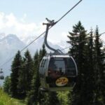 Eine Seilbahn fährt einen Berghang hinauf, umgeben von Kiefern und fernen schneebedeckten Gipfeln unter einem klaren Himmel.