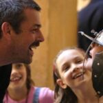 Ein Mann lächelt und spricht mit einem Kind, das einen Metallhelm trägt, während zwei andere Kinder im Hintergrund lächeln.