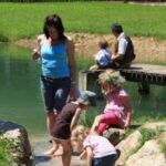 Eine Frau und drei Kinder spielen am Rande eines Teichs, während zwei Personen an einem sonnigen Tag auf einem Holzsteg im Hintergrund sitzen.