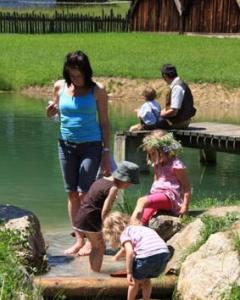 Eine Frau und drei Kinder spielen am Rande eines Teichs, während zwei Personen an einem sonnigen Tag auf einem Holzsteg im Hintergrund sitzen.