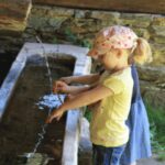 Zwei kleine Kinder stehen nebeneinander und spielen mit dem Wasser, das aus einem Ausguss in ein Steinbecken im Freien fließt.