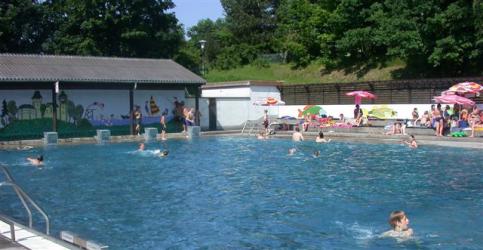 Menschen schwimmen in einem Außenpool, andere sitzen auf Liegestühlen und Handtüchern unter Sonnenschirmen in der Nähe; ein Wandgemälde ist auf einem Gebäude neben dem Pool zu sehen.