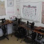 Drei alte Nähmaschinen stehen nebeneinander an einer Wand, die mit bestickten Stoffen und Mustertüchern dekoriert ist.