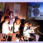 Sieben Personen, teils sitzend, teils stehend, stehen mit Sonnenbrillen auf der Bühne, hinter ihnen sind projizierte Bilder und Zeichen zu sehen.