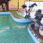 Hallenbad mit einer kleinen Brücke, künstlichen Palmen, Liegestühlen und einigen Sitzgelegenheiten am Beckenrand.