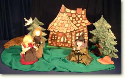 Drei Puppen stehen vor einem Papphaus und Bäumen, die auf einer grünen Stofffläche stehen, mit einer Vogelfigur im Hintergrund.