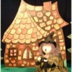 Eine als Hexe verkleidete Puppe steht vor einer Kulisse, die ein mit Süßigkeiten und Keksen dekoriertes Lebkuchenhaus zeigt.