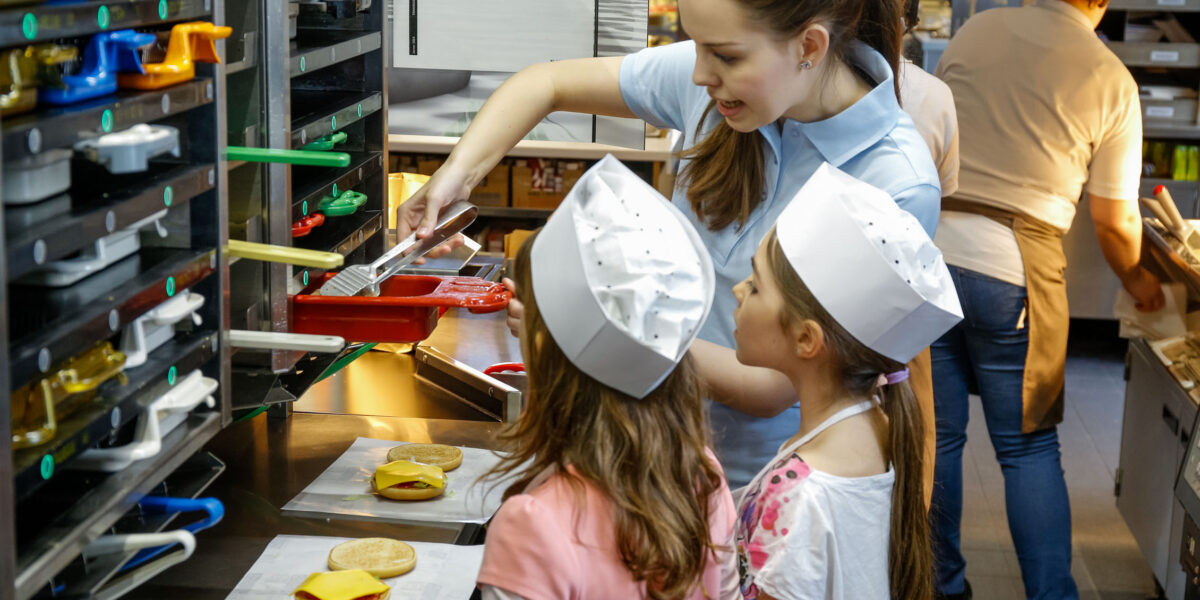 Eine Frau weist zwei Kinder mit Kochmützen an, wie man Burger an der Theke eines Schnellrestaurants zusammenstellt.