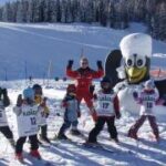 Eine Gruppe von Kindern in nummerierten Westen und ein Skilehrer posieren auf verschneitem Boden neben einer großen Schneemannfigur in einem Skigebiet.