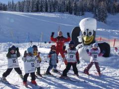 Eine Gruppe von Kindern in nummerierten Westen und ein Skilehrer posieren auf verschneitem Boden neben einer großen Schneemannfigur in einem Skigebiet.