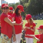 Eine Gruppe von Menschen und Kindern in roten Hemden und Hüten steht an einem sonnigen Tag auf einer Wiese in einem Park zusammen.