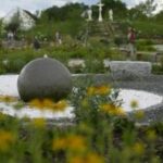 Ein steinerner Kugelbrunnen steht in einem Landschaftsgarten mit gelben Blumen und Steinbänken, im Hintergrund sind ein Kreuz und Statuen zu sehen.