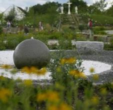Ein steinerner Kugelbrunnen steht in einem Landschaftsgarten mit gelben Blumen und Steinbänken, im Hintergrund sind ein Kreuz und Statuen zu sehen.