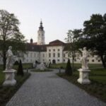 Ein mit Statuen gesäumter Kopfsteinpflasterweg führt zu einem großen weißen historischen Gebäude mit einem zentralen Uhrenturm, das von Bäumen und Gärten umgeben ist.