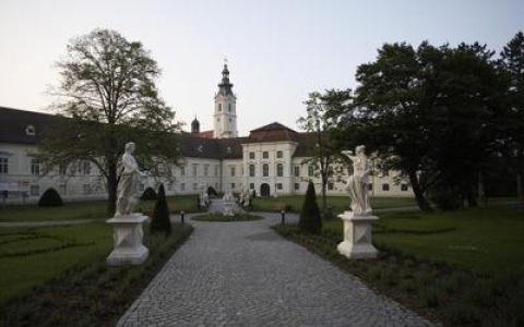 Ein mit Statuen gesäumter Kopfsteinpflasterweg führt zu einem großen weißen historischen Gebäude mit einem zentralen Uhrenturm, das von Bäumen und Gärten umgeben ist.