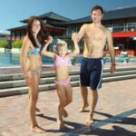 Ein Mann, eine Frau und ein junges Mädchen in Badeanzügen stehen zusammen an einem Freibad, halten sich an den Händen und lächeln in die Kamera.