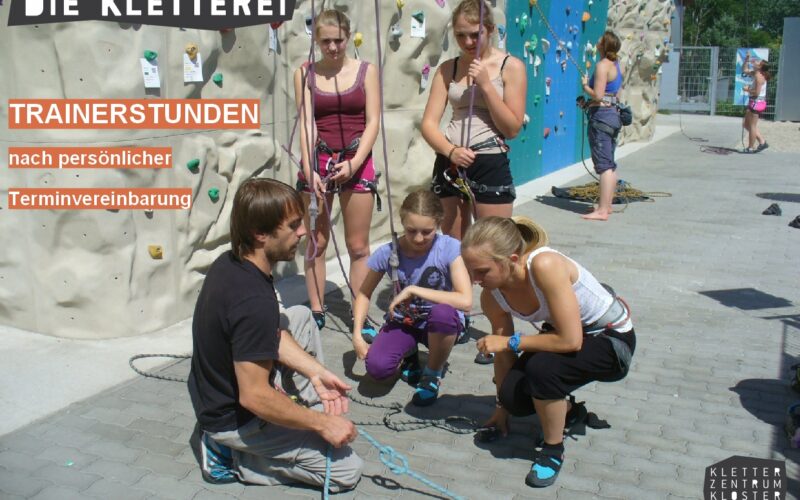 Ein Kletterlehrer demonstriert einer Gruppe von Frauen und Mädchen in einem Kletterzentrum im Freien Seiltechniken, mit Kletterwänden im Hintergrund.