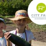 Zwei Kinder mit Hüten halten eine große Zucchini in einem Garten, in der oberen rechten Ecke ist das Logo der City Farm Schönbrunn zu sehen.