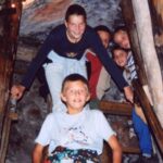Fünf Kinder steigen eine schmale Holztreppe in einer Höhle oder einem Tunnel hinunter, lächeln und halten sich an den Geländern fest.