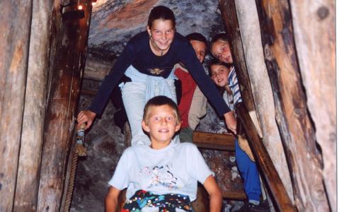 Fünf Kinder steigen eine schmale Holztreppe in einer Höhle oder einem Tunnel hinunter, lächeln und halten sich an den Geländern fest.