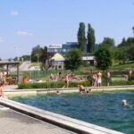 Menschen schwimmen und entspannen sich an einem sonnigen Tag in einem Freibad, während im Hintergrund Gebäude und Bäume zu sehen sind.