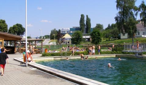 Menschen schwimmen und entspannen sich an einem sonnigen Tag in einem Freibad, während im Hintergrund Gebäude und Bäume zu sehen sind.