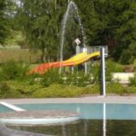 Freibad mit kreisförmigen Plattformen, einem Springbrunnen, der Wasser versprüht, und einer gelb-orangenen Rutsche im Hintergrund, umgeben von Grün und Bäumen.