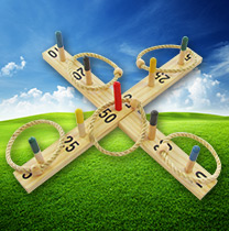 Ein hölzernes Ringwurfspiel mit nummerierten Pflöcken und Seilringen wird auf grünem Gras unter einem blauen Himmel mit vereinzelten Wolken platziert.