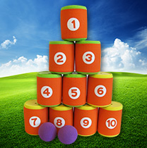 Zehn orangefarbene und grüne Schaumstoffdosen, nummeriert von 1 bis 10, sind in einer Pyramide auf Gras gestapelt, mit zwei lila Bällen davor, vor einem blauen Himmel mit Wolken.