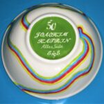 Eine weiße Schale mit bunten Wellenlinien, grünem Boden und weißer Schrift "50 Joachim Katrin Alles Gute 666" auf blauem Hintergrund.