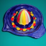 Eine farbenfrohe Zitruspresse aus Keramik steht auf einer tealfarbenen Oberfläche. Der Entsaftungskegel ist mit vertikalen Streifen in Rot, Gelb, Blau und Weiß versehen.