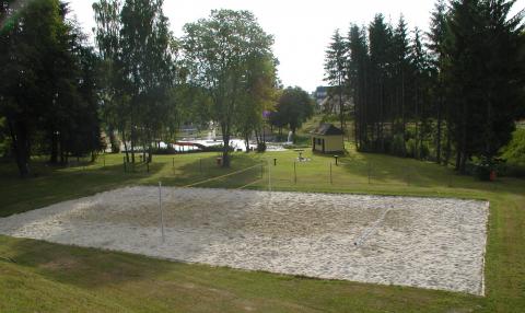 Im Freien ist ein Sandvolleyballfeld auf einer Wiese aufgebaut, umgeben von Bäumen und einigen Gebäuden im Hintergrund.