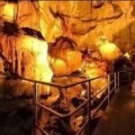 Ein gut beleuchteter Metallsteg führt durch das Innere einer Höhle mit orangefarbenen und braunen Felswänden.