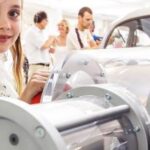 Ein junges Mädchen steht bei einer Indoor-Veranstaltung neben einer transparenten Maschine oder einem Exponat, während im Hintergrund Menschen und ein Auto zu sehen sind.