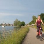Zwei Personen fahren mit ihren Fahrrädern auf einem gepflasterten Weg neben einem See, der von Gras und Bäumen gesäumt ist, während im Hintergrund ein Dorf zu sehen ist.
