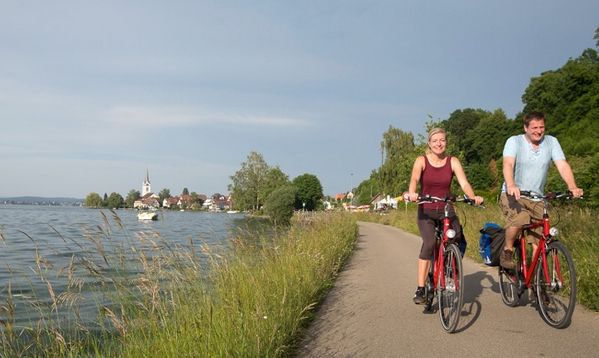 Zwei Personen fahren mit ihren Fahrrädern auf einem gepflasterten Weg neben einem See, der von Gras und Bäumen gesäumt ist, während im Hintergrund ein Dorf zu sehen ist.