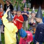 Eine Gruppe von Kindern mit kegelförmigen Hüten versammelt sich im Freien, einige stehend, andere hockend, mit einem erwachsenen Betreuer im Hintergrund.