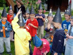 Eine Gruppe von Kindern mit kegelförmigen Hüten versammelt sich im Freien, einige stehend, andere hockend, mit einem erwachsenen Betreuer im Hintergrund.