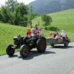 Eine Gruppe von Kindern fährt auf einem Anhänger, der von einem Traktor gezogen wird, eine Landstraße entlang, mit grünen Hügeln und einem Spaziergänger in der Nähe.
