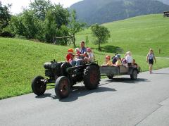 Eine Gruppe von Kindern fährt auf einem Anhänger, der von einem Traktor gezogen wird, eine Landstraße entlang, mit grünen Hügeln und einem Spaziergänger in der Nähe.