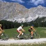 Drei Personen fahren mit ihren Fahrrädern auf einem Schotterweg durch eine grasbewachsene Gegend mit großen Bergen und einem Wald im Hintergrund unter einem blauen Himmel.
