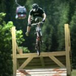 Eine Person mit Helm fährt mit einem Mountainbike von einer Holzrampe mit einer Seilbahn und Bäumen im Hintergrund.