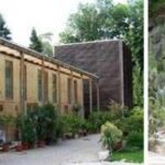 Ein langgestrecktes Gewächshaus mit hohen Fenstern ist von Topfpflanzen umgeben; rechts daneben befindet sich ein steiniger Gartenbereich mit verschiedenen Kakteen und Sukkulenten.