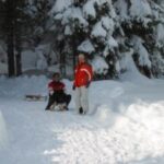 Zwei Personen mit einem kleinen Schlitten stehen auf einem schneebedeckten Weg, der von schneebedeckten Bäumen in einem Waldgebiet umgeben ist.