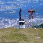 Eine Seilbahn fährt einen grasbewachsenen Berghang hinauf, während im Hintergrund eine Stadt und eine Landschaft zu sehen sind.