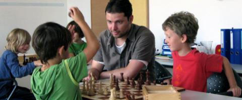 Ein Erwachsener und drei Kinder sitzen an einem Tisch und spielen Schach. Der Erwachsene schaut auf das Brett, während ein Kind eine Figur zieht und ein anderes Kind zusieht.