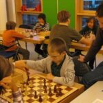 Kinder spielen Schach in einem Klassenzimmer, während ein Erwachsener sie beaufsichtigt; Schachbretter und Stühle sind auf Tischen angeordnet.