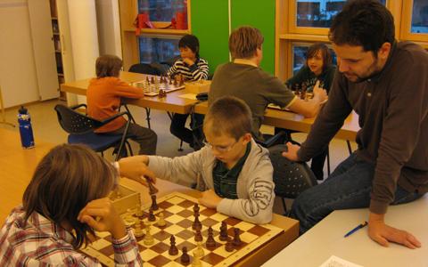 Kinder spielen Schach in einem Klassenzimmer, während ein Erwachsener sie beaufsichtigt; Schachbretter und Stühle sind auf Tischen angeordnet.
