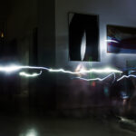 Ein lang belichtetes Foto fängt eine helle, wellenförmige Lichtlinie in einem schummrigen Raum mit verschwommenen Figuren und Kunstwerken an den Wänden ein.