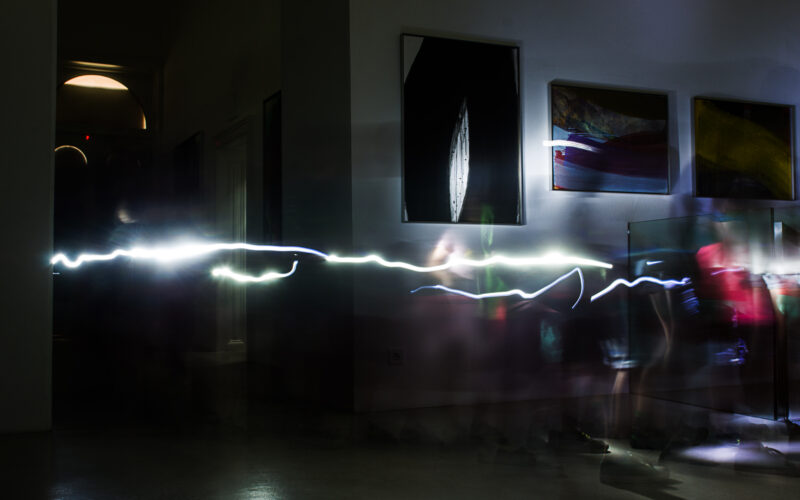 Ein lang belichtetes Foto fängt eine helle, wellenförmige Lichtlinie in einem schummrigen Raum mit verschwommenen Figuren und Kunstwerken an den Wänden ein.